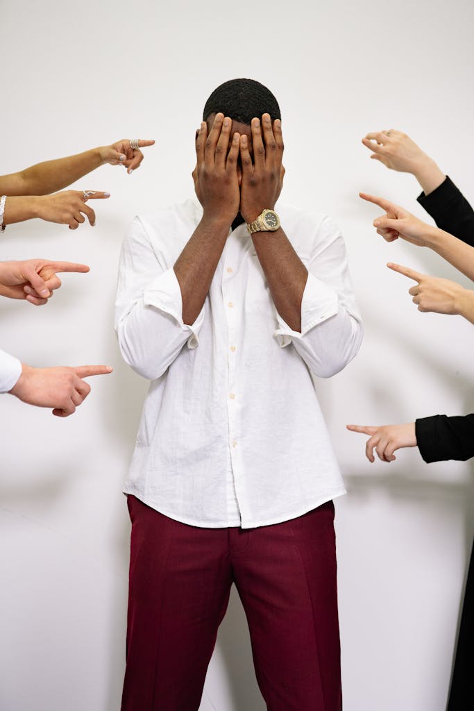 A man in distress covers his face surrounded by multiple pointing fingers.