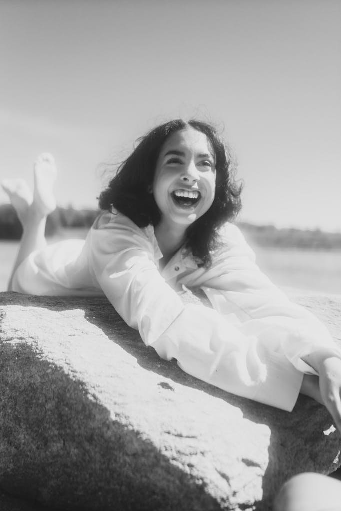 A cheerful adult woman with a big smile reclining on a rock by a serene lake, capturing a moment of pure happiness.